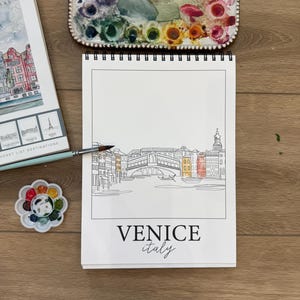 May include: A sketchbook with a line drawing of Venice, Italy, featuring the Rialto Bridge, with the text "VENICE italy". A watercolor palette and paintbrushes are nearby. Other travel-themed items are also present.