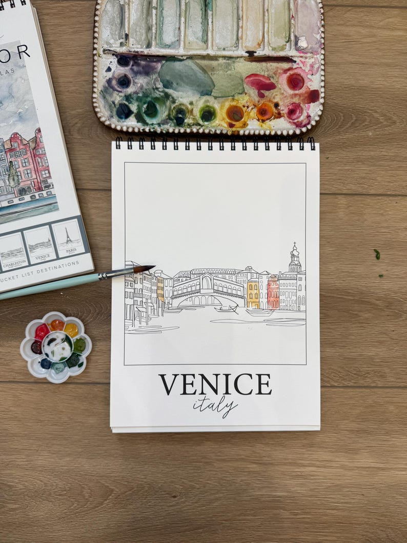 May include: A sketchbook with a line drawing of Venice, Italy, featuring the Rialto Bridge, with the text "VENICE italy". A watercolor palette and paintbrushes are nearby. Other travel-themed items are also present.