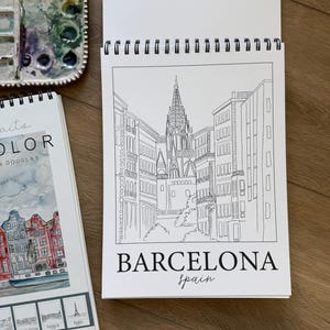 May include: A spiral-bound sketchbook open to a black and white line drawing of a Barcelona cityscape, with the word "BARCELONA" and "spain" below. A watercolor palette and a travel-themed book are also visible.