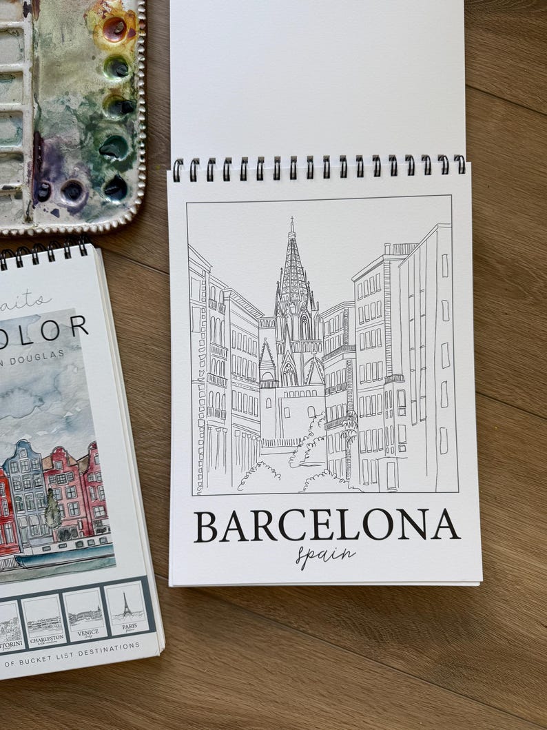 May include: A spiral-bound sketchbook open to a black and white line drawing of a Barcelona cityscape, with the word "BARCELONA" and "spain" below. A watercolor palette and a travel-themed book are also visible.