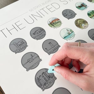 May include: A white print titled "50 State Bucket List" of the United States, with illustrations of each state. A light blue tool is scratching off the Illinois state illustration. The print is on a white surface.