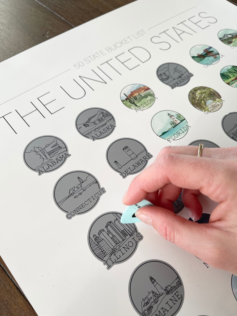 May include: A white print titled "50 State Bucket List" of the United States, with illustrations of each state. A light blue tool is scratching off the Illinois state illustration. The print is on a white surface.