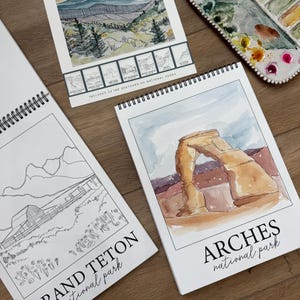 May include: Watercolor workbooks featuring national park sketches. One workbook is open to a watercolor painting of Arches National Park. Another workbook shows a black and white sketch of Grand Teton National Park. A watercolor palette is also visible.