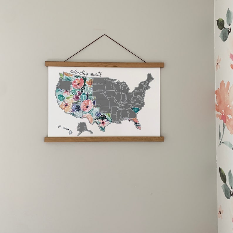 May include: A framed map of the United States with a floral design overlay. The map is gray with state names printed in white. The floral design is in shades of pink, purple, blue, and green. The frame is made of wood and has a light brown finish. The text "adventure awaits" is printed at the top of the map.