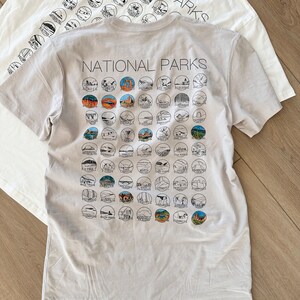 Puede incluir: Una camiseta gris claro con el texto "NATIONAL PARKS" impreso en la parte delantera. La parte posterior de la camiseta presenta una cuadr&iacute;cula de 50 c&iacute;rculos peque&ntilde;os, cada uno con una ilustraci&oacute;n en blanco y negro de un parque nacional diferente.