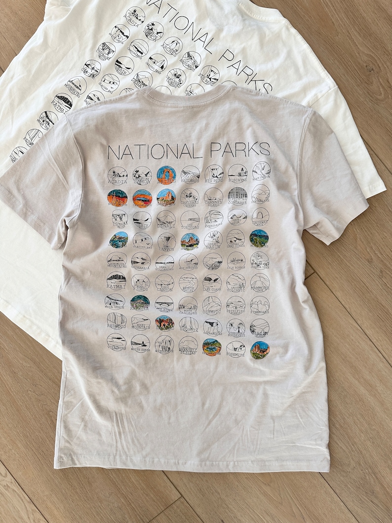 Puede incluir: Una camiseta gris claro con el texto "NATIONAL PARKS" impreso en la parte delantera. La parte posterior de la camiseta presenta una cuadr&iacute;cula de 50 c&iacute;rculos peque&ntilde;os, cada uno con una ilustraci&oacute;n en blanco y negro de un parque nacional diferente.