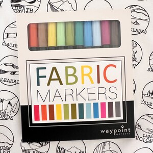 Puede incluir: Un juego de marcadores para tela en una caja blanca con las palabras "FABRIC MARKERS" impresas en la parte delantera. La caja est&aacute; abierta y muestra los marcadores en una variedad de colores, incluyendo rojo, naranja, amarillo, verde, azul, morado y marr&oacute;n. La caja tambi&eacute;n tiene las palabras "waypoint wanders" impresas en la parte inferior.