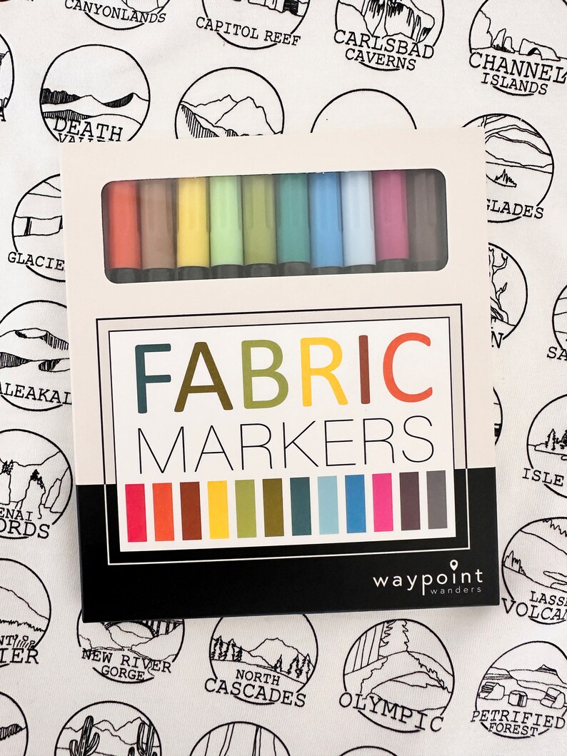 Puede incluir: Un juego de marcadores para tela en una caja blanca con las palabras "FABRIC MARKERS" impresas en la parte delantera. La caja est&aacute; abierta y muestra los marcadores en una variedad de colores, incluyendo rojo, naranja, amarillo, verde, azul, morado y marr&oacute;n. La caja tambi&eacute;n tiene las palabras "waypoint wanders" impresas en la parte inferior.
