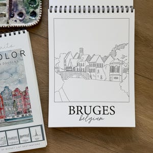 May include: A white spiral-bound sketchbook with a black and white line drawing of Bruges, Belgium, and the word "BRUGES" below. A watercolor palette and a book with watercolor illustrations are also visible.