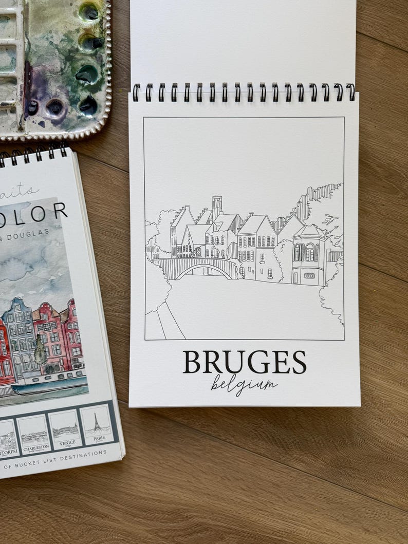 May include: A white spiral-bound sketchbook with a black and white line drawing of Bruges, Belgium, and the word "BRUGES" below. A watercolor palette and a book with watercolor illustrations are also visible.