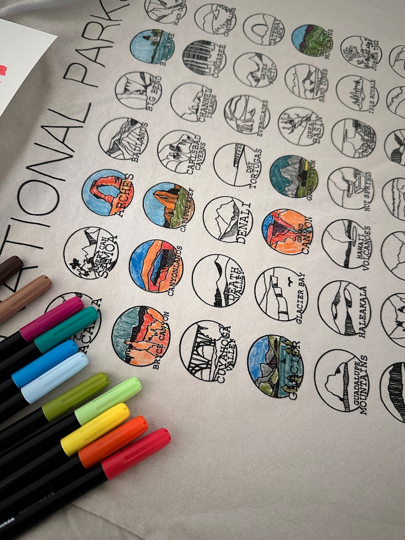 Puede incluir: Una camiseta blanca con un dise&ntilde;o de 30 parques nacionales diferentes de los Estados Unidos. Cada parque est&aacute; representado por un c&iacute;rculo con un contorno negro y una ilustraci&oacute;n a color de un hito o caracter&iacute;stica del parque. La camiseta est&aacute; rodeada de marcadores de colores.