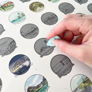 May include: A scratch-off map of the United States with illustrations of each state. The map features a hand holding a tool to scratch off the foil layer of the Massachusetts state illustration. The map includes state names and illustrations.