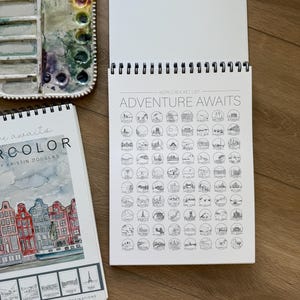 May include: An open spiral-bound sketchbook with the text "ADVENTURE AWAITS" and a world bucket list. A watercolor palette with various colors and a sketchbook with the text "watercolor" are also visible.