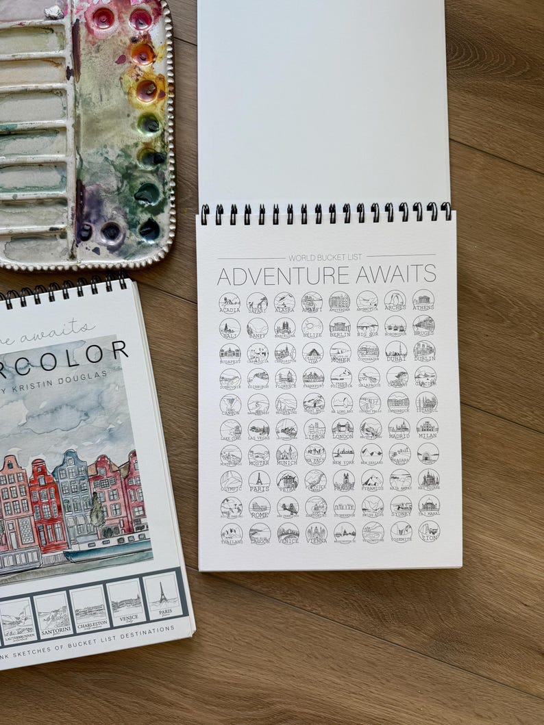 May include: An open spiral-bound sketchbook with the text "ADVENTURE AWAITS" and a world bucket list. A watercolor palette with various colors and a sketchbook with the text "watercolor" are also visible.