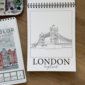 May include: A spiral-bound sketchbook featuring a black and white line drawing of the Tower Bridge in London. The words "LONDON england" are printed below the illustration. A watercolor palette and a book with destination illustrations are also visible.
