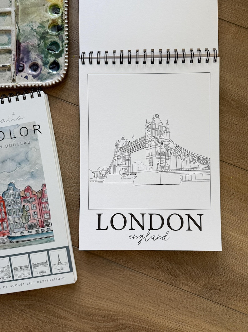 May include: A spiral-bound sketchbook featuring a black and white line drawing of the Tower Bridge in London. The words "LONDON england" are printed below the illustration. A watercolor palette and a book with destination illustrations are also visible.
