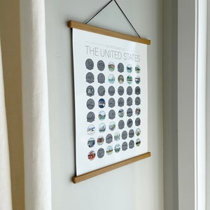May include: A framed poster titled "The United States 50 State Bucket List" hangs on a wall. The poster features illustrations of landmarks and attractions from each state. The frame is made of light-colored wood, and the poster is suspended by a black cord.