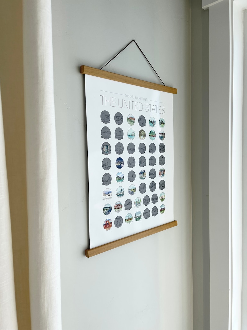 May include: A framed poster titled "The United States 50 State Bucket List" hangs on a wall. The poster features illustrations of landmarks and attractions from each state. The frame is made of light-colored wood, and the poster is suspended by a black cord.