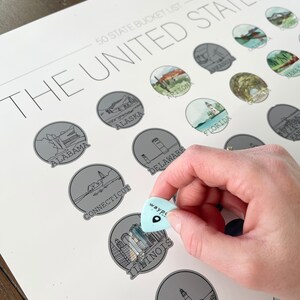May include: A 50 State Bucket List print featuring illustrations of U.S. states. A light blue guitar pick with the word "wayp" is held over the Illinois illustration. The print includes state names and illustrations in a circular design.