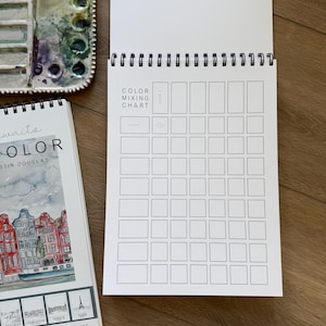 May include: A watercolor artist's setup featuring a spiral-bound color mixing chart, a watercolor palette with various paint colors, and a sketchbook titled "Color" with destination illustrations. The chart is open, ready for use.