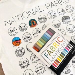 Puede incluir: Una camiseta blanca con un contorno negro de varios parques nacionales. La camiseta est&aacute; puesta plana y una caja de marcadores de tela est&aacute; encima de la camiseta. Los marcadores son de varios colores, incluyendo rojo, naranja, amarillo, verde, azul, morado y negro. El texto "NATIONAL PARKS" est&aacute; impreso en la parte superior de la camiseta.
