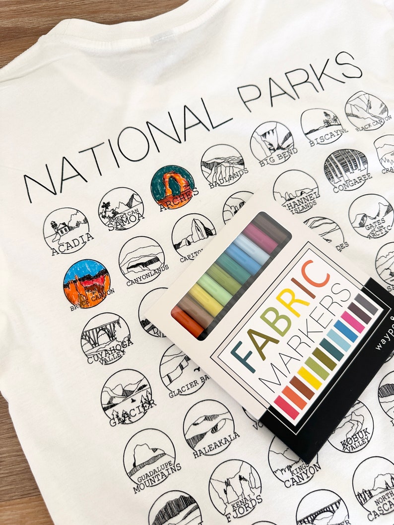 Puede incluir: Una camiseta blanca con un contorno negro de varios parques nacionales. La camiseta est&aacute; puesta plana y una caja de marcadores de tela est&aacute; encima de la camiseta. Los marcadores son de varios colores, incluyendo rojo, naranja, amarillo, verde, azul, morado y negro. El texto "NATIONAL PARKS" est&aacute; impreso en la parte superior de la camiseta.