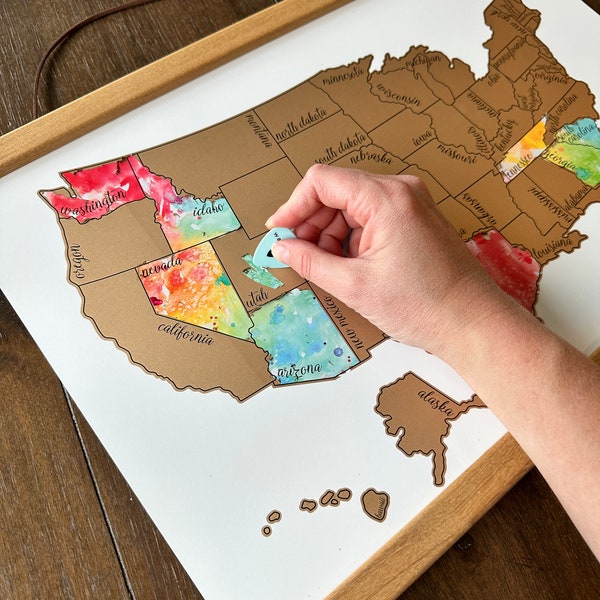 May include: A framed United States map with scratch-off state outlines. The map features watercolor-style state colors. A person is holding a small, blue, teardrop-shaped object, presumably to scratch off a state. The map is mounted on a wooden frame.