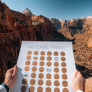 May include: A white poster with the text "NATIONAL PARKS" at the top. The poster is divided into a grid of 6 rows and 7 columns, with each square containing a small, circular image of a national park. The poster is being held up in front of a scenic view of a canyon.