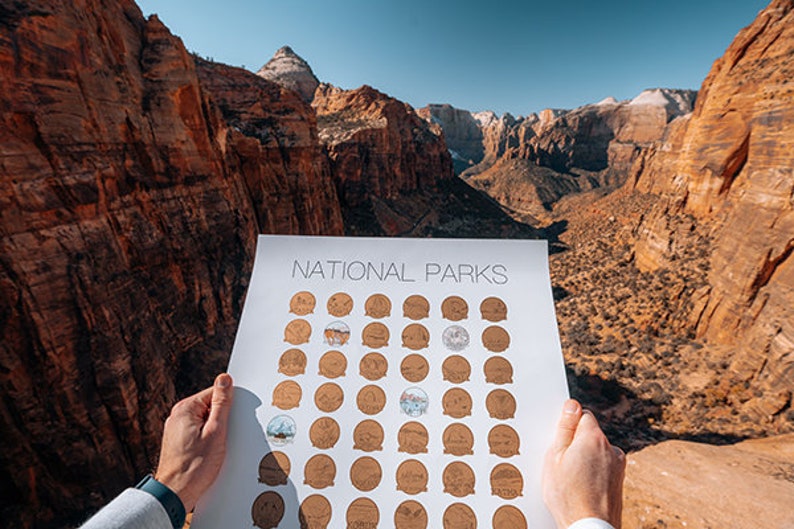 May include: A white poster with the text "NATIONAL PARKS" at the top. The poster is divided into a grid of 6 rows and 7 columns, with each square containing a small, circular image of a national park. The poster is being held up in front of a scenic view of a canyon.