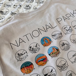 Puede incluir: Camiseta blanca con una impresi&oacute;n en negro de los nombres de varios Parques Nacionales de los Estados Unidos. Los nombres est&aacute;n dispuestos en un c&iacute;rculo alrededor de las palabras "National Parks".
