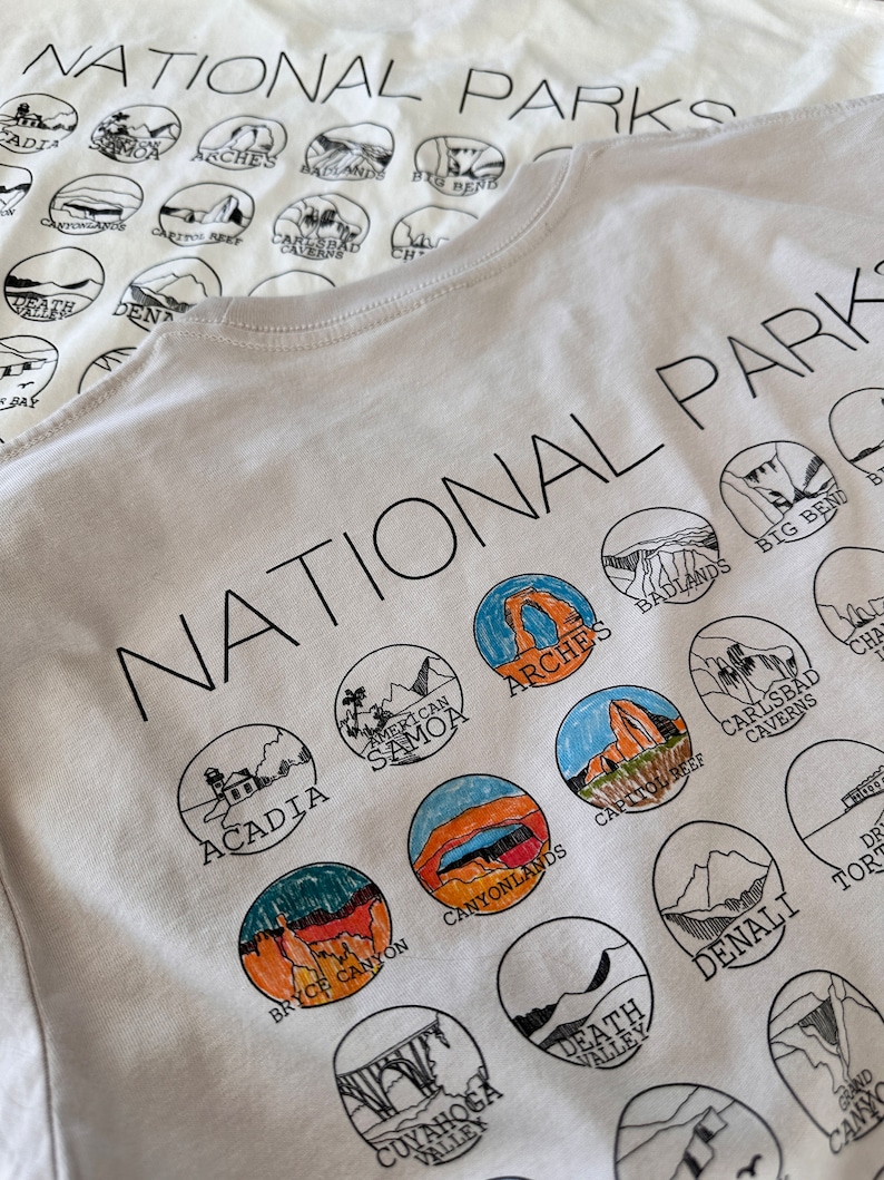 Puede incluir: Camiseta blanca con una impresi&oacute;n en negro de los nombres de varios Parques Nacionales de los Estados Unidos. Los nombres est&aacute;n dispuestos en un c&iacute;rculo alrededor de las palabras "National Parks".