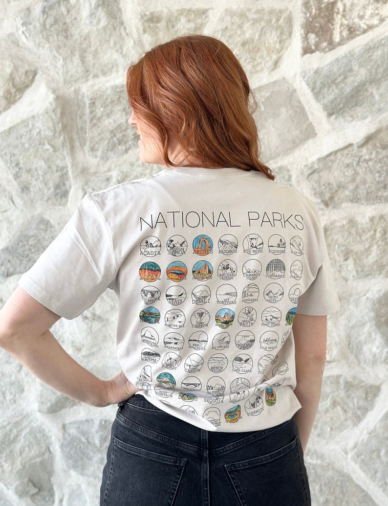 Puede incluir: Una camiseta blanca con el texto "NATIONAL PARKS" impreso en la espalda. La camiseta presenta un dise&ntilde;o de varios logotipos de parques nacionales, incluidos Arches, Yosemite y Yellowstone.