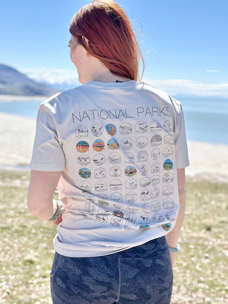 Puede incluir: Una camiseta gris claro con el texto "National Parks" impreso en la espalda. La camiseta presenta un dise&ntilde;o de 50 parques nacionales diferentes en los Estados Unidos, cada uno representado por un c&iacute;rculo con un icono &uacute;nico.