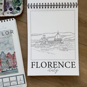 May include: A spiral-bound sketchbook featuring a black and white line drawing of Florence, Italy, with the text "FLORENCE italy" at the bottom. A watercolor palette and a travel-themed sketchbook are also visible.