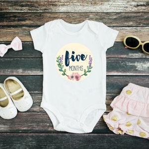 May include: White baby onesie with a yellow circle and the text "five months" in navy blue lettering. The circle is decorated with green leaves and pink flowers. The onesie is laid out on a wooden surface with a pink bow, white shoes, pink and gold polka dot shorts, and gold sunglasses.