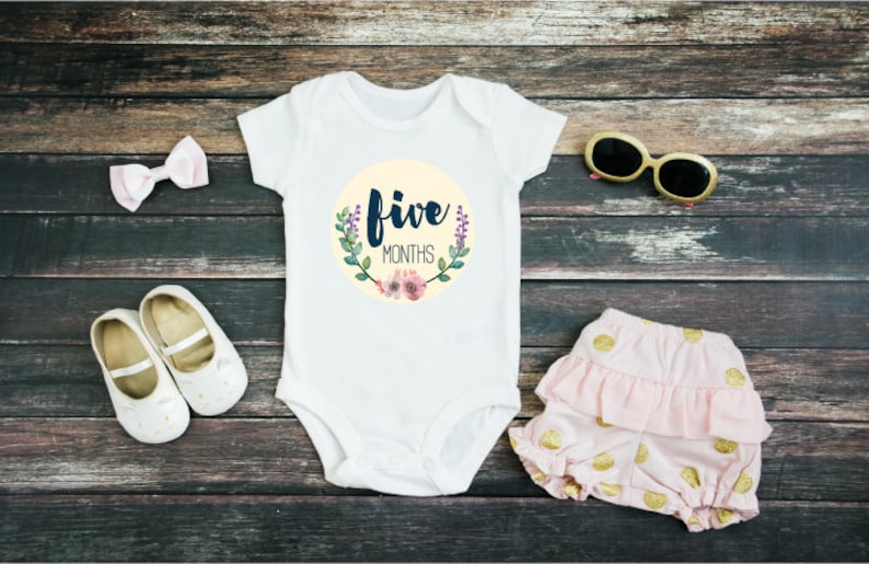 May include: White baby onesie with a yellow circle and the text "five months" in navy blue lettering. The circle is decorated with green leaves and pink flowers. The onesie is laid out on a wooden surface with a pink bow, white shoes, pink and gold polka dot shorts, and gold sunglasses.