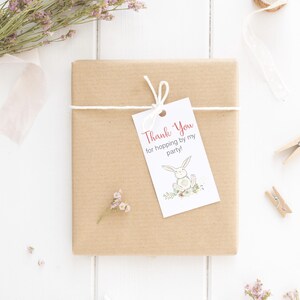 Pu&ograve; includere: Un regalo avvolto in carta marrone con un'etichetta bianca che dice "Thank You for hopping by my party!" e un'illustrazione di un coniglietto. Il regalo &egrave; legato con uno spago bianco e decorato con fiori secchi e un nastro.