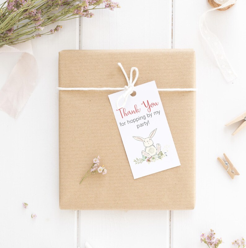 Pu&ograve; includere: Un regalo avvolto in carta marrone con un'etichetta bianca che dice "Thank You for hopping by my party!" e un'illustrazione di un coniglietto. Il regalo &egrave; legato con uno spago bianco e decorato con fiori secchi e un nastro.