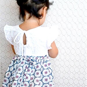 May include: A young girl wearing a white dress with a floral print skirt. The dress has a lace bodice and a tie at the back. The girl has her hair pulled back in two pigtails.