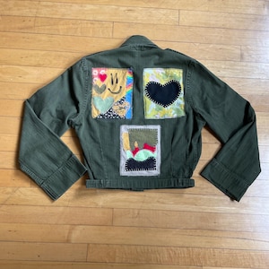 May include: Olive green jacket with three colorful patches on the back. The patches feature a smiley face, a black heart, and an abstract design. The jacket has long sleeves and a button-up front.