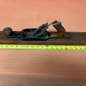 May include: A vintage wooden hand plane with a black metal blade and handle. The plane is approximately 22 inches long. The wooden body is a dark brown color, and the handle shows signs of wear. A yellow measuring tape is placed next to the plane.