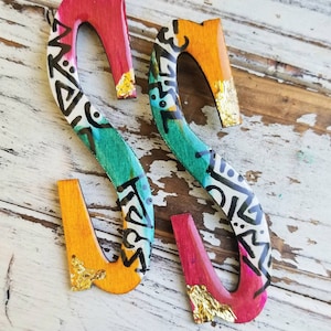 May include: Two wooden letter "S" cutouts, hand-painted with vibrant colors and patterns. The letters feature a mix of orange, teal, and pink hues, accented with black tribal designs and gold leaf details. Decorative home decor.