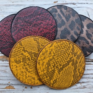 May include: A collection of round coasters with various animal print designs. The coasters feature patterns like snake skin in red and yellow, and leopard print in brown and black. Each coaster has a black border.