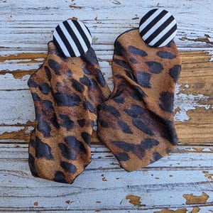 May include: A pair of fabric earrings with a leopard print design. The earrings are brown and black with a button top that has black and white stripes. The earrings are displayed on a distressed, white-painted wooden surface.
