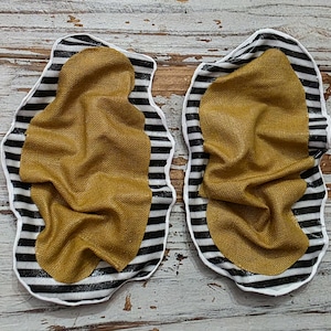 May include: Two decorative fabric pieces. Each has a gold-colored, textured center, surrounded by a black and white striped border. The edges of the pieces are wavy, creating a unique shape. The background is a light-colored, wooden surface.