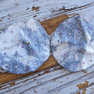 May include: Two denim-like fabric appliques, each with a petal shape, are adorned with scattered iridescent rhinestones. The fabric has a blue and white tie-dye effect. The appliques are displayed on a weathered wooden surface.