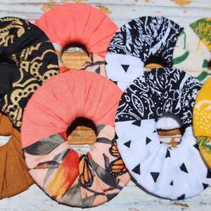 May include: Six fabric hair scrunchies in various patterns and colors. The scrunchies are made of different fabrics, including black and gold paisley, peach, white with black triangles, and yellow with green and white.