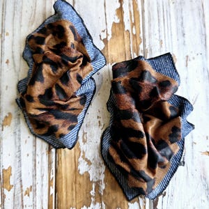May include: Two fabric hair accessories. Each features a brown and black leopard print fabric, layered over a blue denim-like fabric. The edges are finished with a black trim. The items are displayed on a distressed, white-painted wooden surface.