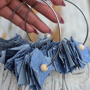 May include: Large silver hoop earrings with a denim fringe. Each earring features a cluster of blue denim fabric pieces and a small wooden bead accent. The earrings are displayed against a light-colored wooden surface.