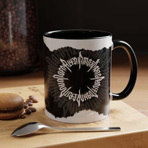 May include: Black and white ceramic mug with a black handle and interior. The mug features a circular design with stylized text in a gothic font. The mug sits on a wooden surface with a spoon and a pastry.